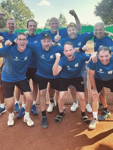 Gruppe von neun jubelnden Männern in blauen T-Shirts, stehen an einem Tennisplatz hinter einem Netz.