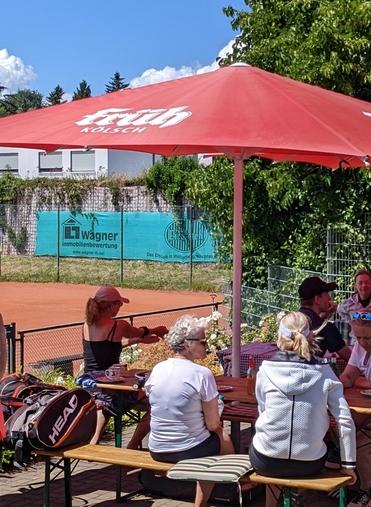Gruppengespräch unter einem roten Sonnenschirm auf einem Tennisplatz, umgeben von grüner Vegetation und blauen Himmel.