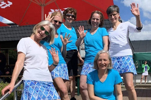Sechs Frauen in sportlicher Kleidung stehen auf Treppenstufen und winken in die Kamera. Sonniger Tag mit Regenschirm im Hintergrund.