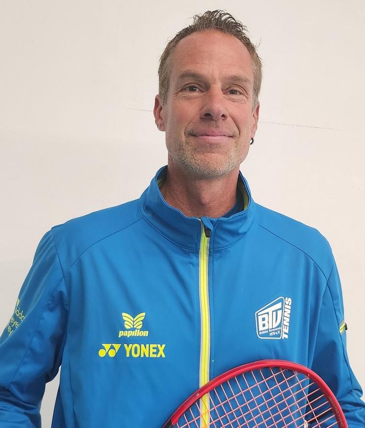 Mann in blauer Sportjacke mit Tennisschläger lächelt vor weißer Wand.