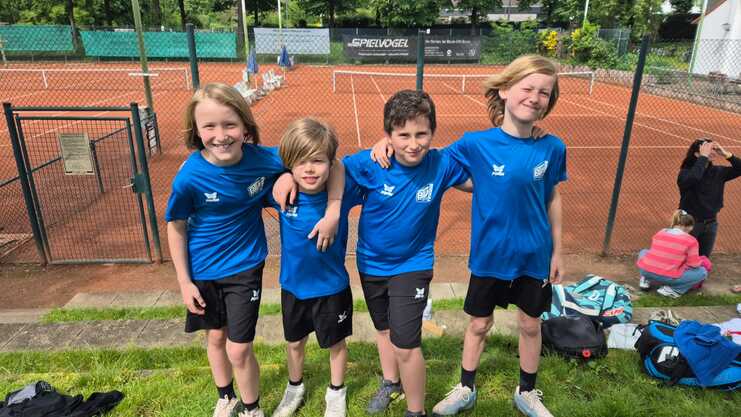 Vier Kinder in blauen T-Shirts stehen lächelnd und umarmend vor einem Tennisplatz mit Sandbelag.