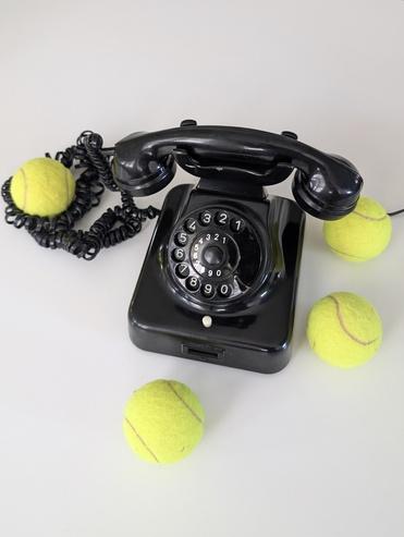 Schwarz-weißes Telefon mit Wählscheibe, umgeben von drei gelben Tennisbällen auf einem Tisch.