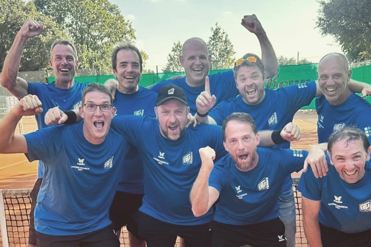 Gruppe von neun jubelnden Männern in blauen T-Shirts, stehen an einem Tennisplatz hinter einem Netz.