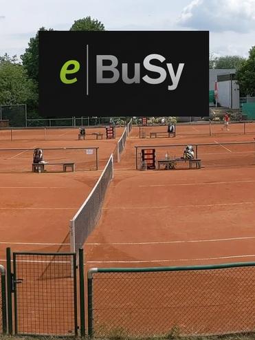 Panoramaaufnahme eines Tennisplatzes mit Spielern, Umrandungen und Gebäuden im Hintergrund unter einem bewölkten Himmel.
