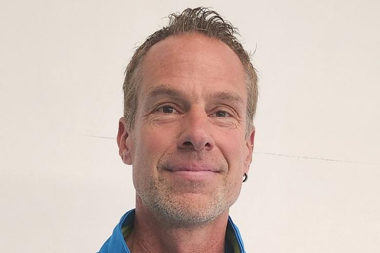 Mann in blauer Sportjacke mit Tennisschläger lächelt vor weißer Wand.