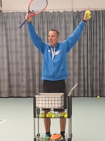 Mann in blauer Jacke hebt Schläger und Ball, steht vor einem Wagen mit Tennisbällen auf einem Tennisplatz.