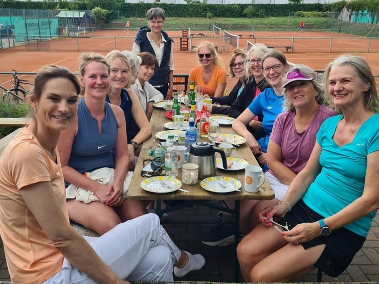Gruppe von zwölf Frauen sitzt an einem Tisch mit Snacks und Getränken, im Hintergrund Tennisplätze und blauer Himmel.