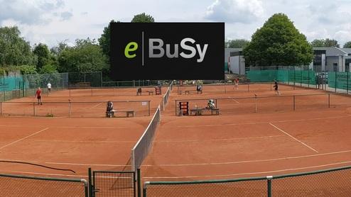 Panoramaaufnahme eines Tennisplatzes mit Spielern, Umrandungen und Gebäuden im Hintergrund unter einem bewölkten Himmel.