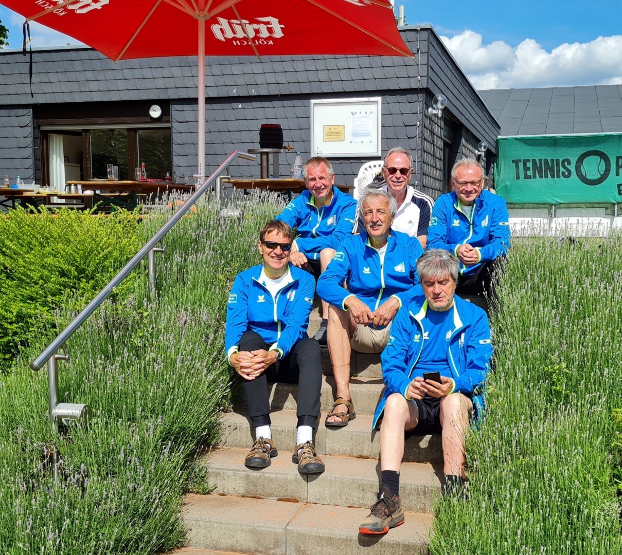 Sechs Personen in blauen Jacken sitzen auf einer Treppe, umgeben von Lavendel, vor einem Tennisplatz.