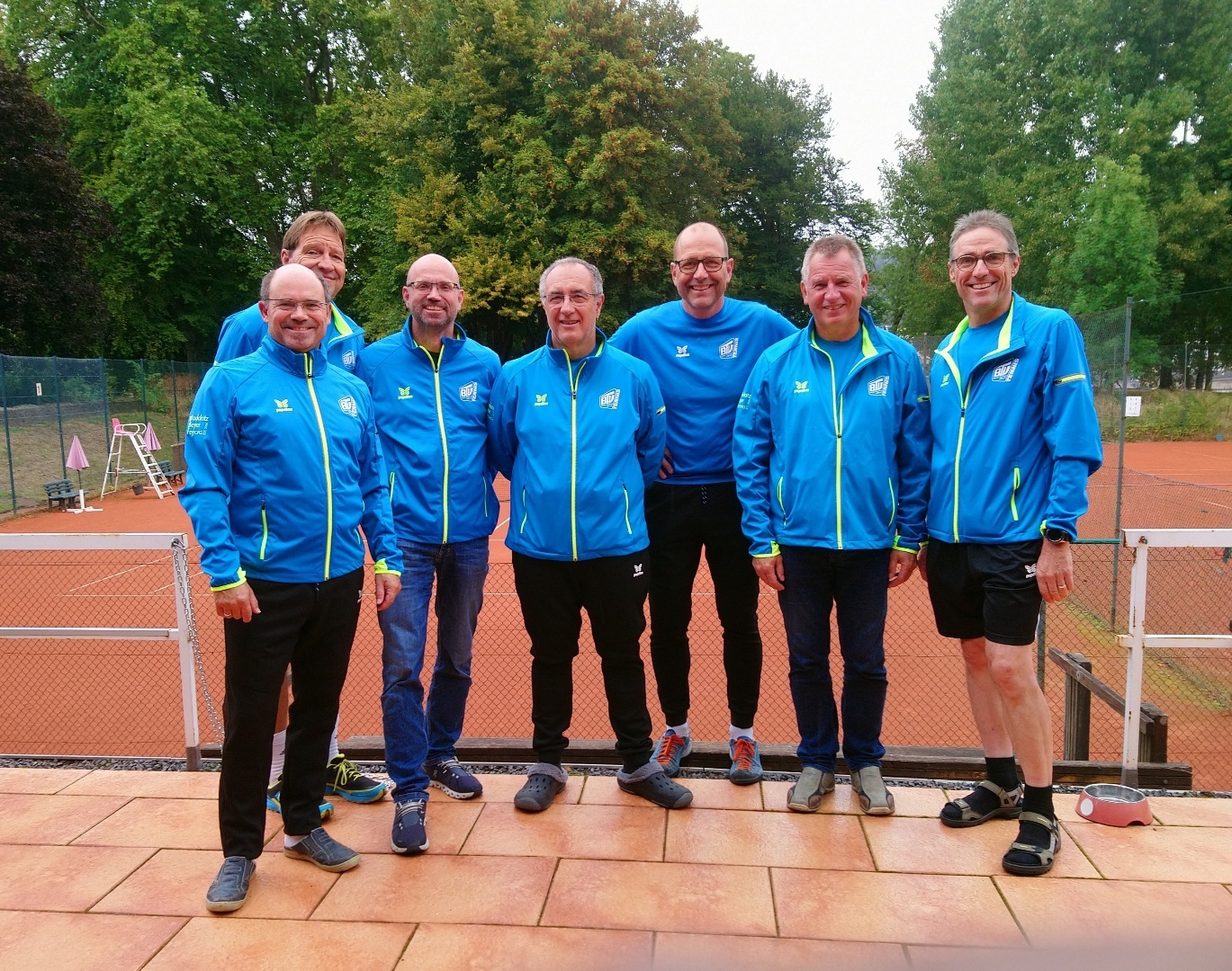 Gruppe von Männern in blauen Sportjacken steht auf einem Tennisplatz mit Bäumen im Hintergrund.