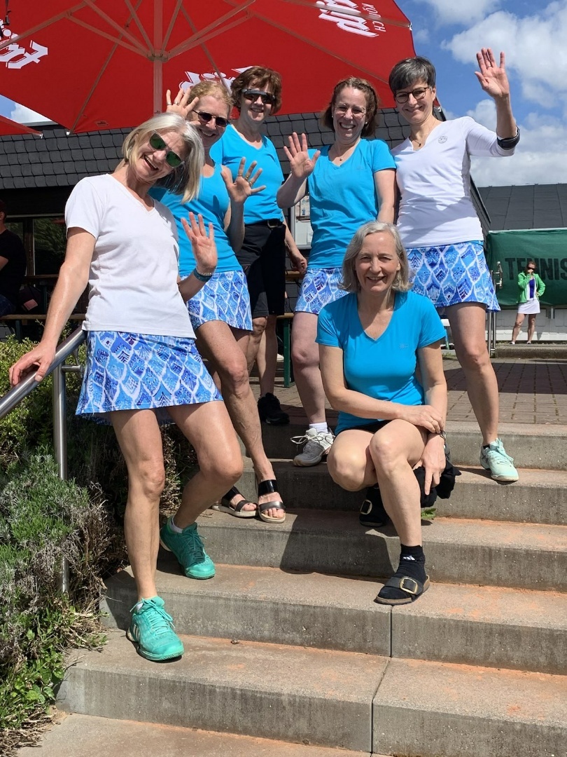 Sechs Frauen in sportlicher Kleidung stehen auf Treppenstufen und winken in die Kamera. Sonniger Tag mit Regenschirm im Hintergrund.