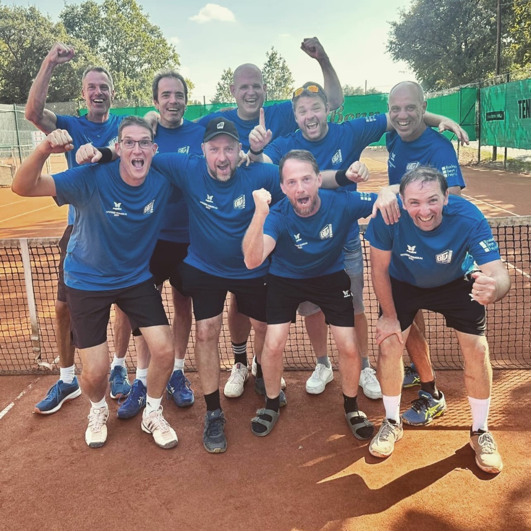 Gruppe von neun jubelnden Männern in blauen T-Shirts, stehen an einem Tennisplatz hinter einem Netz.
