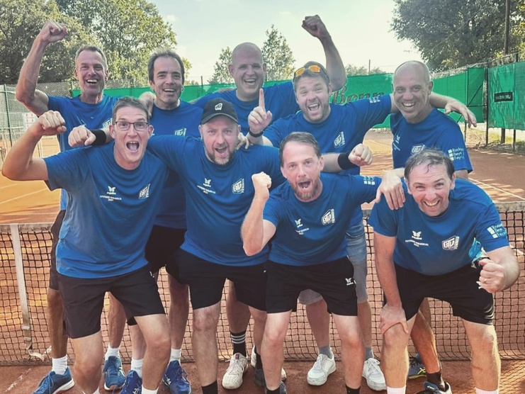 Gruppe von neun jubelnden Männern in blauen T-Shirts, stehen an einem Tennisplatz hinter einem Netz.