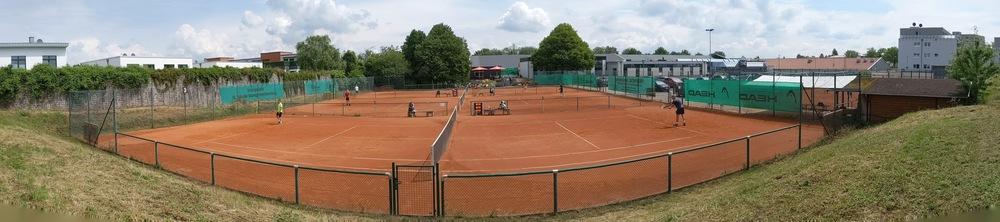 Panoramaaufnahme eines Tennisplatzes mit mehreren Spielern auf rotem Sand und umliegenden Gebäuden und Bäumen.