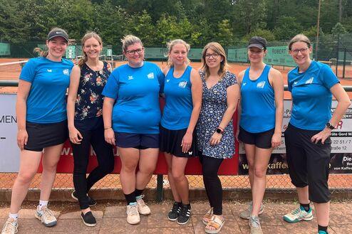 Gruppenfoto von sieben Frauen in blauen Trikots vor einem Tennisplatz mit Bäumen im Hintergrund.