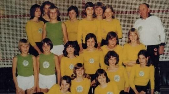 Gruppenfoto von Mädchen in gelben und grünen Trikots mit einem Trainer, in einer Sporthalle positioniert.