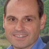 Ein Mann mit kurzen, braunen Haaren und einem freundlichen Lächeln vor einer grünen Landschaft.