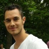 Ein junger Mann mit kurzem, dunklem Haar trägt ein weißes T-Shirt und lächelt vor einer grünen Baumkulisse.