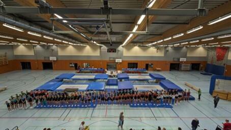 Blick auf eine Sporthalle mit mehreren Trampolinen und einer Gruppe von Sportlern, die sich in Formation aufstellen.