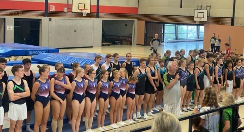 Gruppe von Turnerinnen und Turnern in Sportanzügen stehen aufgereiht in einer Turnhalle, während ein Trainer spricht.