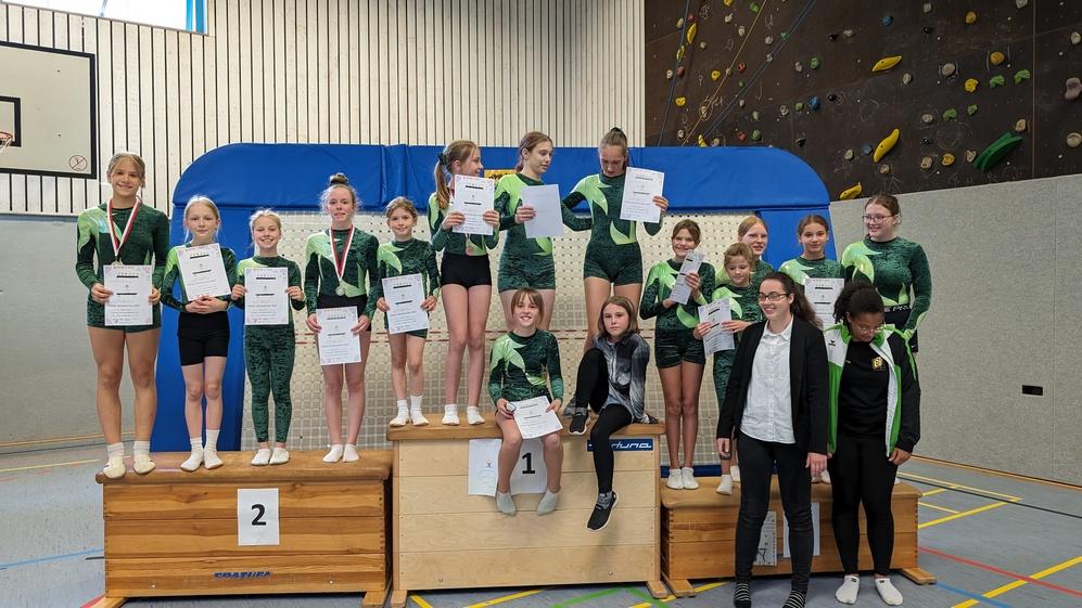 Gruppenbild von Turnerinnen in grünen Outfits auf einem Siegertreppchen, halten Urkunden und Medaillen nach Wettkampf.