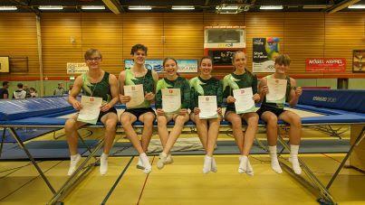 Sechs Trampolinsportler in Sportkleidung sitzen auf einem Trampolin und halten Urkunden in der Hand.