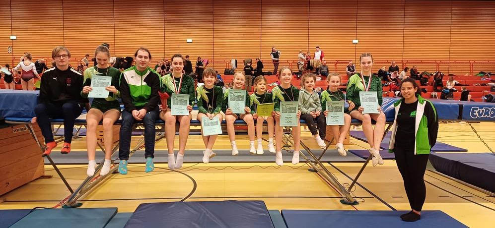 Gruppe von Sportlern in Gymnastikanzügen hält Urkunden und Medaillen, sitzt auf Geräten in einer Turnhalle.