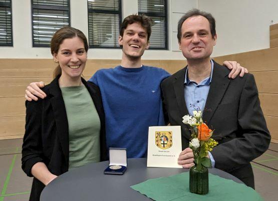 Drei Personen stehen an einem Tisch, halten eine Auszeichnung und lächeln, mit einer Blumenvase im Vordergrund.