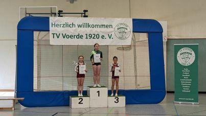 Drei Sportlerinnen auf einem Siegertreppchen, mit Urkunden, vor einer Begrüßungsbanner des TV Voerde 1920 e. V.