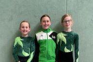 Drei junge Turnerinnen stehen nebeneinander in Sportkleidung mit grünem Blattdesign vor einer grauen Wand.