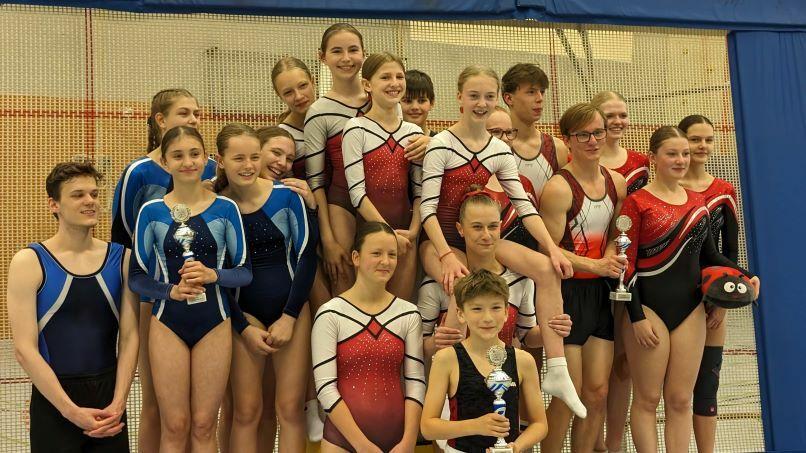 Gruppe von Turnerinnen und Turnern in Wettkampfanzügen, posierend mit Pokalen auf einem Gymnastikgelände.