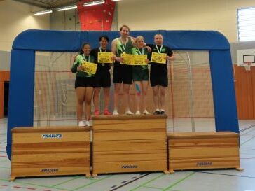 Fünf Sportlerinnen stehen auf einem Siegertreppchen und halten Urkunden in der Sporthalle.