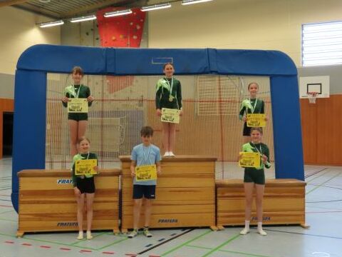 Fünf Kinder stehen auf Podesten in einer Sporthalle und halten Urkunden in den Händen. Sie tragen Sportkleidung und Medaillen.