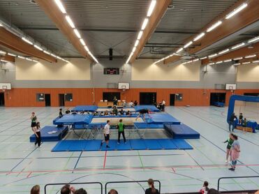 Blick auf eine Trampolinhalle mit mehreren Trampolinen und Sportlern in Aktion, Zuschauer auf Tribüne im Vordergrund.