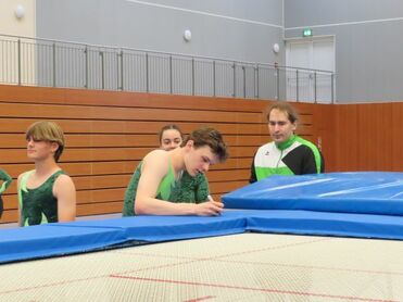 Vier Trampolinsportler in Training, einer zeigt auf sein Handy, während andere zuschauen. Trampolin und Sporthalle im Hintergrund.