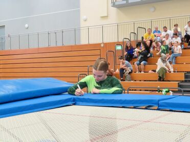 Mädchen in grüner Sportkleidung schreibt auf dem Rand eines Trampolins, im Hintergrund Zuschauer auf Tribünen.
