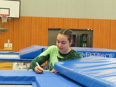 Jugendliche in grünem Sportanzug schreibt mit einem Marker auf einer blauen Trampolinplatte in der Turnhalle.