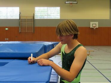 Junge mit blonden Haaren schreibt mit einem Marker auf eine blaue Turnmatte in einer Sporthalle.