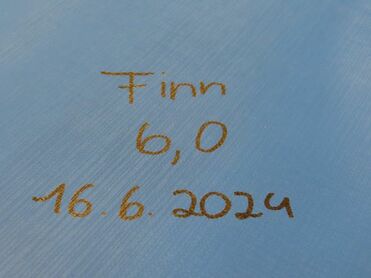 Beige Schrift auf blauer Oberfläche mit den Worten: „Finn 6,0 16.6.2024“.