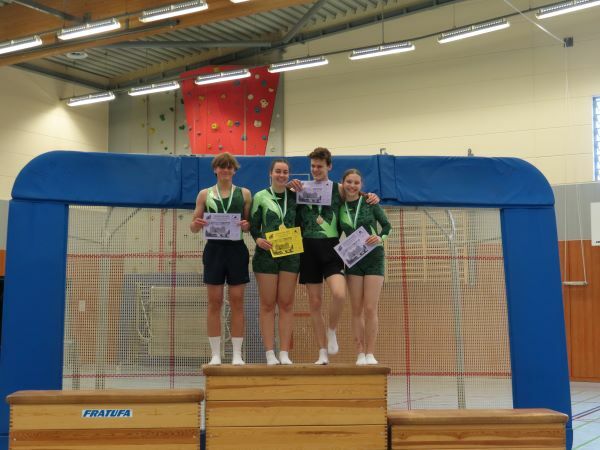 Vier Athleten stehen auf einem Siegerpodest in der Turnhalle, halten Urkunden und Medaillen in der Hand.