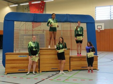 Vier sportlich gekleidete Frauen stehen auf einem Podium mit Medaillen, während eine weitere Person daneben steht.