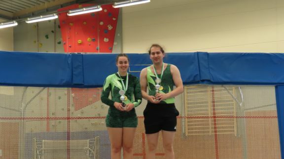 Zwei Personen mit Medaillen stehen vor einem Trampolin in einem Sportzentrum. Die Wand dahinter ist mit Klettergriffen dekoriert.