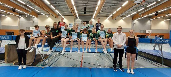 Gruppe von Gymnastikern mit Urkunden, die auf Trampolinen in einer Sporthalle posieren.
