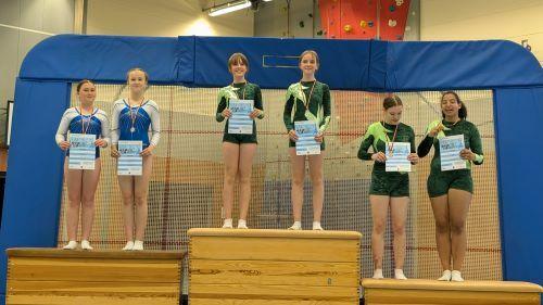 Sechs junge Turnerinnen stehen auf dem Siegerpodest, halten ihre Urkunden und tragen Gymnastikanzüge in verschiedenen Farben.