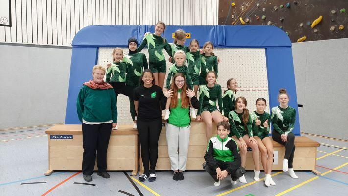Gruppe von Gymnastinnen und Trainern in grünen Übungen auf einer Gymnastikbank in einer Sporthalle.