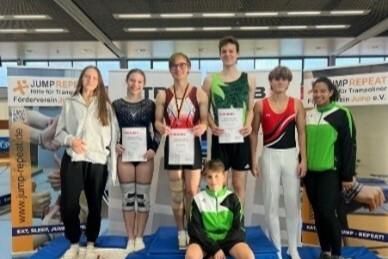 Sechse Athleten stehen mit Urkunden auf dem Siegertreppchen, umgeben von Hintergrundbanner für Trampolinsport.