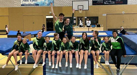Gruppe von 12 Turnerinnen und Turnern in grünen Trikots auf einem Trampolin mit jubelnden Posen im Turnsaal.