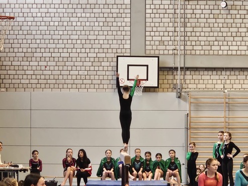 Turnerin in schwarzem Anzug balanciert auf einem Schwebebalken, w&auml;hrend Zuschauer und Richter applaudieren.