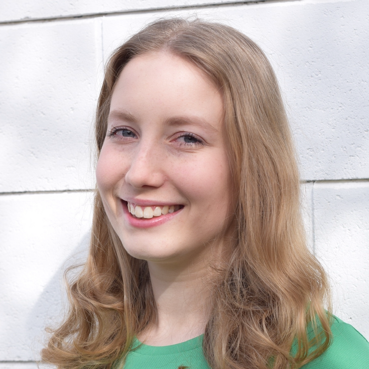 L&auml;chelnde junge Frau mit langen, lockigen Haaren in einem gr&uuml;nen Shirt vor einer wei&szlig;en Wand.