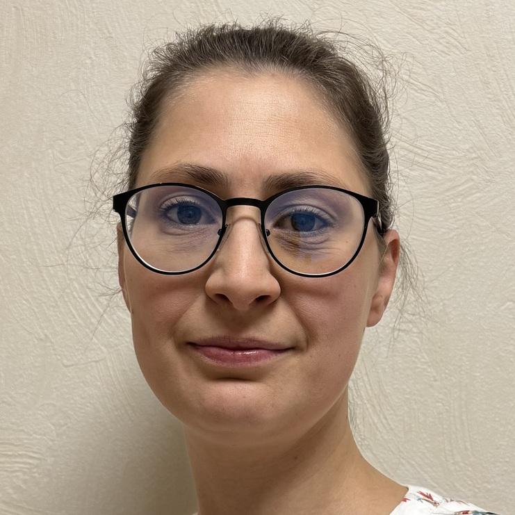 Portrait einer person mit Brille, die vor einer strukturierten Wand steht, trägt ein blütenverziertes Oberteil.